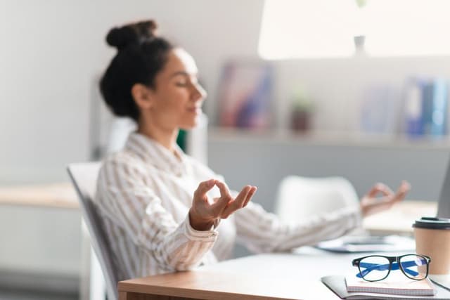 A woman is sitting at a desk with her eyes closed and hands gently raised in a meditative or calming gesture, suggesting a moment of relaxation or mindfulness. On the desk, there is a disposable coffee cup and a pair of glasses resting on a notebook, indicating a break from work or study. The environment appears tranquil with soft natural lighting, contributing to the serene atmosphere the woman seems to be embracing.