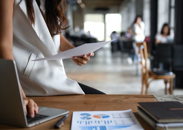 A person is seated at a wooden table with papers and a laptop, reading a document, in an office or coworking space.
