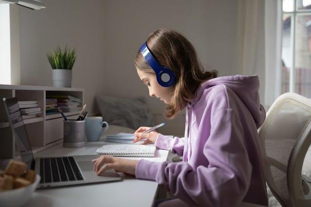 A young person is sitting at a desk wearing a purple hoodie and blue headphones, concentrating on writing in a notebook. In front of them is a laptop computer, suggesting they may be engaged in studying or doing homework. On the desk, there is also a mug and a small container holding pens, adding to the studious setting. The room is well-lit with natural light coming from a window out of view, and there is a shelf with books and a potted plant, creating a calm and organized environment.