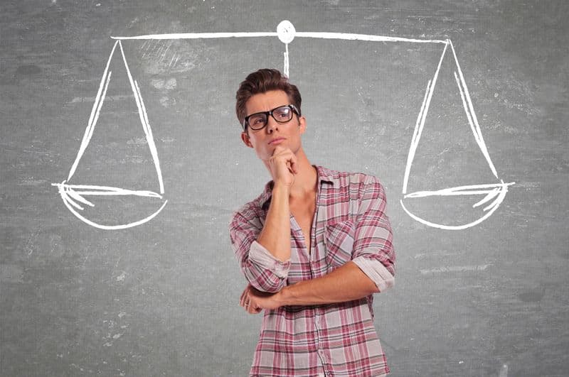 A pensive man with glasses stands in front of a chalkboard with a drawn scale, suggesting contemplation or decision-making.